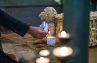 Eine Hand zündet eine Kerze an, die neben einem Teddy und einem Korb steht.