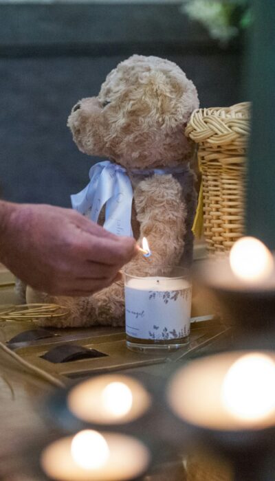 Eine Hand zündet eine Kerze an, die neben einem Teddy und einem Korb steht.