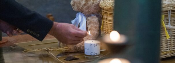 Eine Hand zündet eine Kerze an, die neben einem Teddy und einem Korb steht.