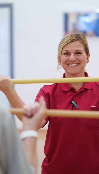 Eine Physiotherapeutin zeigt einem älteren Mann mit einem Stock eine körperliche Übung.