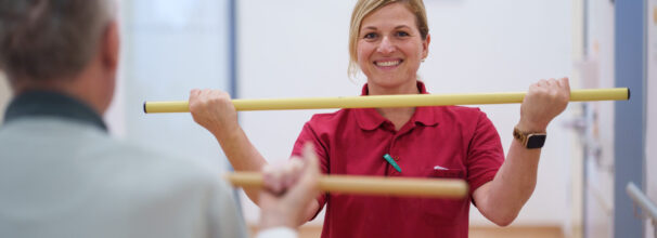 Eine Physiotherapeutin zeigt einem älteren Mann mit einem Stock eine körperliche Übung.