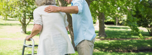 Zwei ältere Personen laufen über eine Wiese mit Bäumen, wobei die weiblich gelesene Person eine Gehhilfe nutzt und die männlich gelesene Person unterstützt.