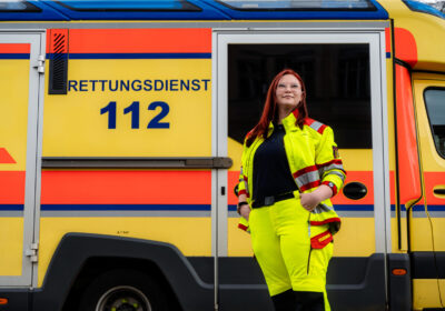 Eine weiblich gelesene Frau posiert vor einem Krankenwagen.