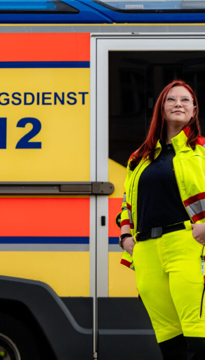 Eine weiblich gelesene Frau posiert vor einem Krankenwagen.